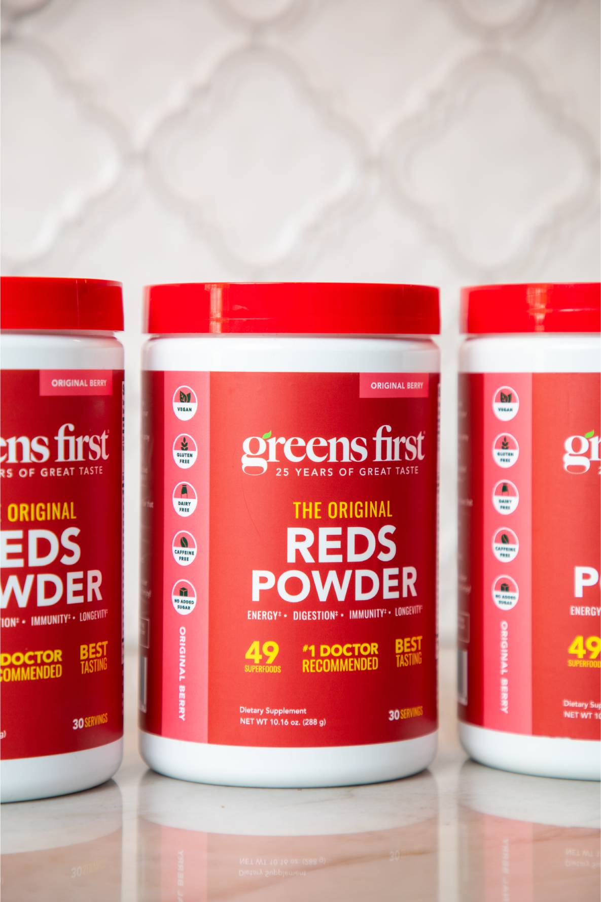Three containers of Greens First Reds Powder on a white surface with a patterned wall in the background.