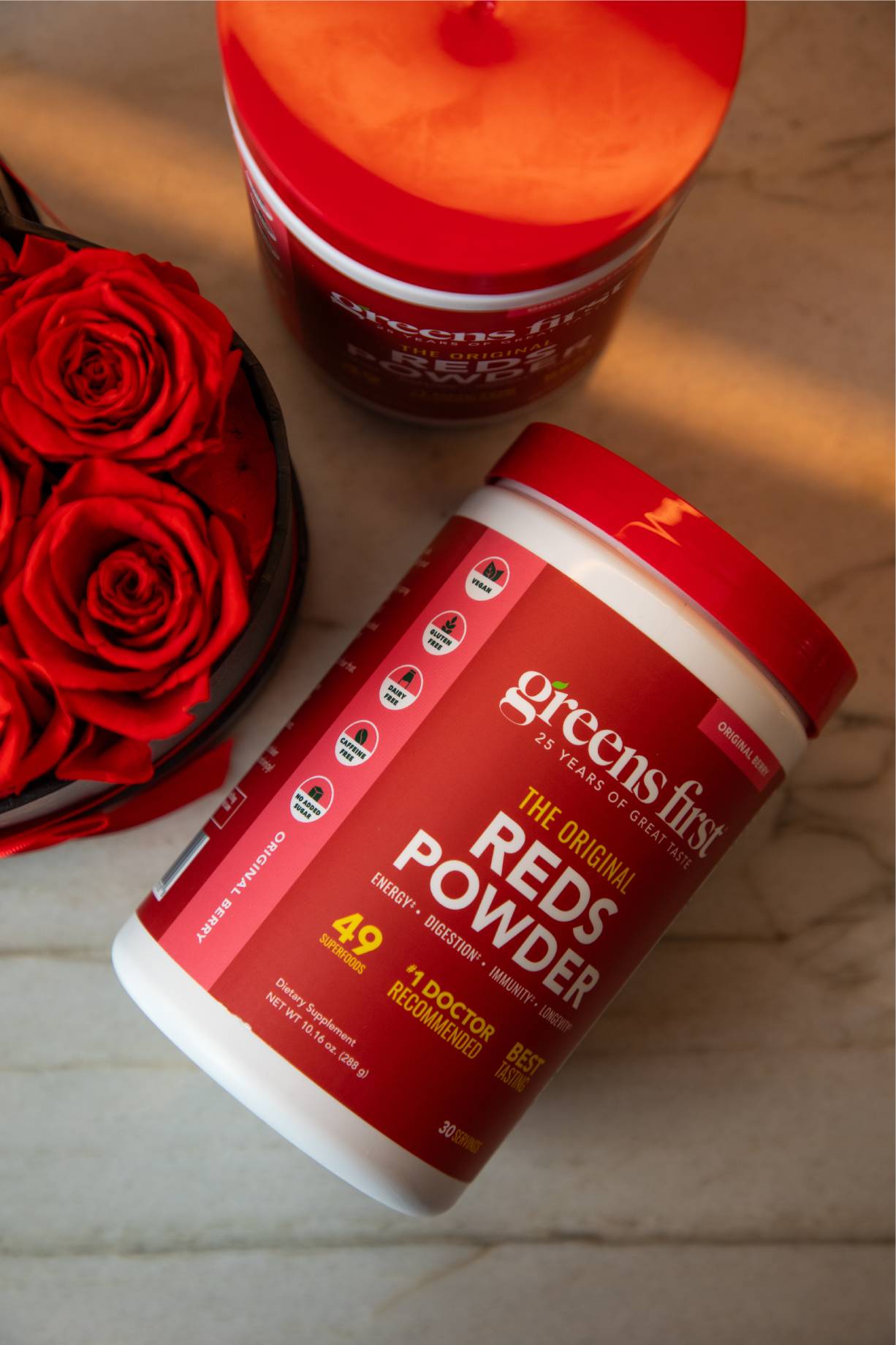 Container of Greens First Reds Powder with red roses on a marble surface