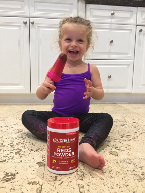 Child holding a red berry popsicle beside a container of Greens First Original Reds Powder